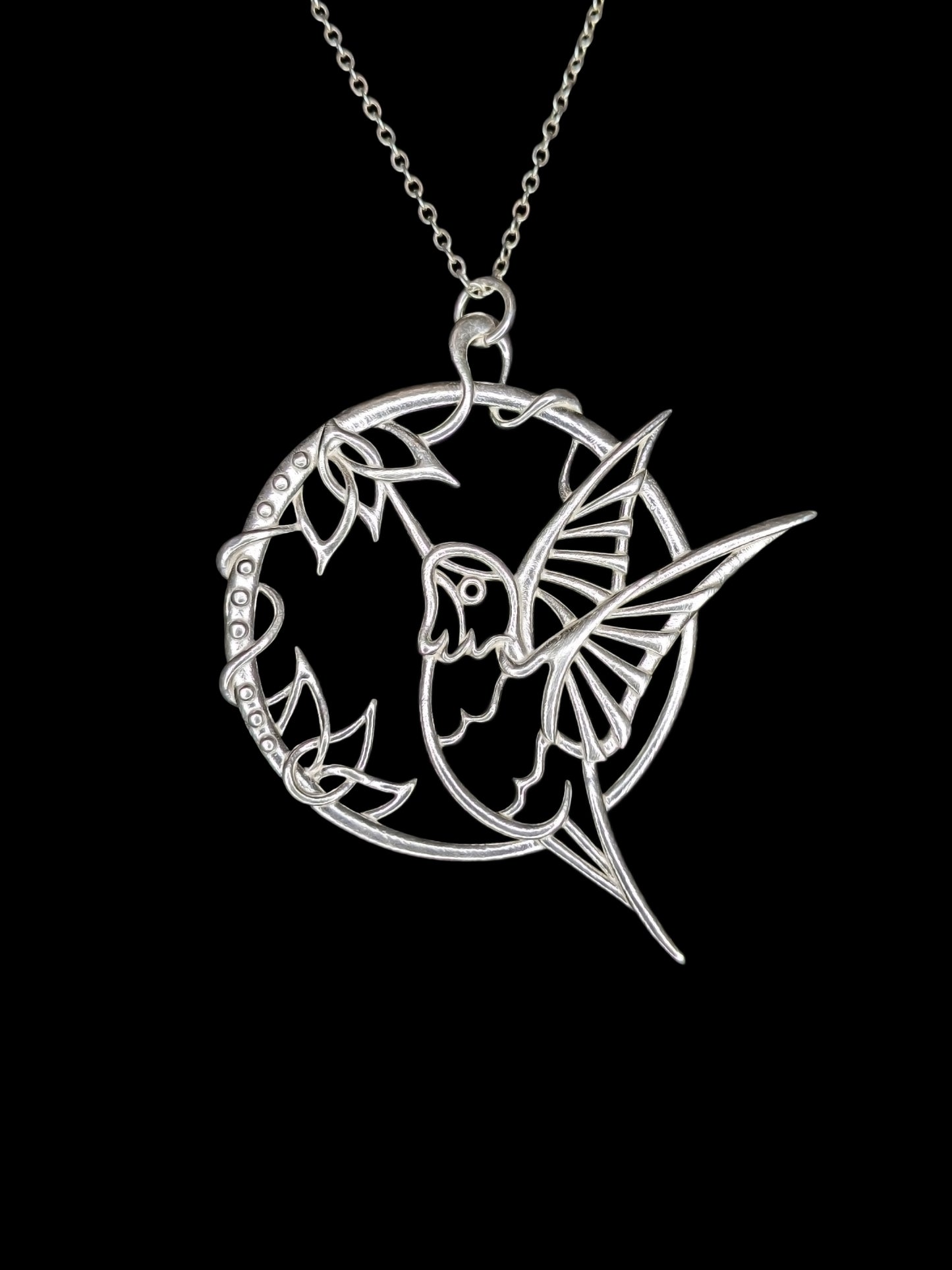 A circular pendant shows a hummingbird in flight drinking from intertwined flowers. The wings and tail extend past the circular frame for a more dynamic shape.