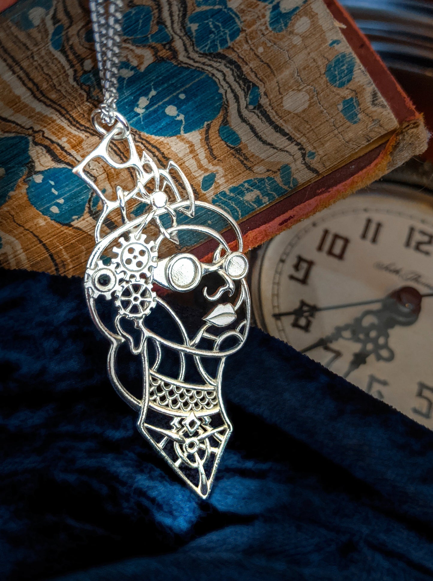 The silver Victoria pendant against blue velvet, a book, and a clock face.